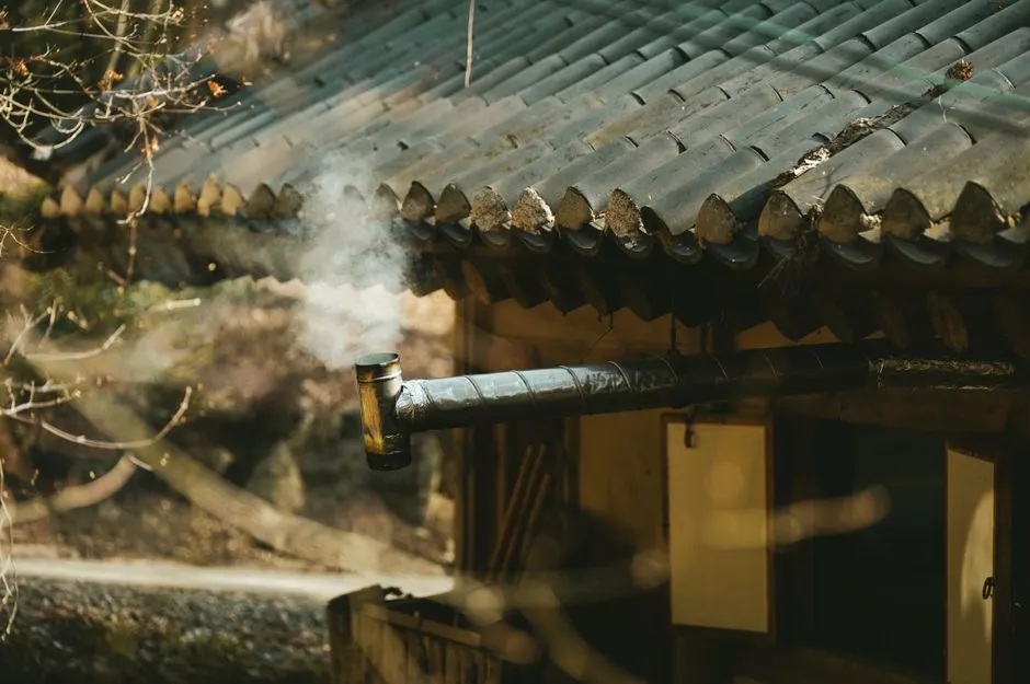 A rustic chimney releasing smoke on a traditional rural house roof, captured during daylight.