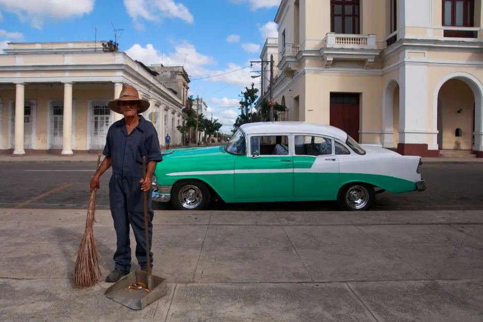A vintage car parked on a street next to a street cleaner in a historic town setting.