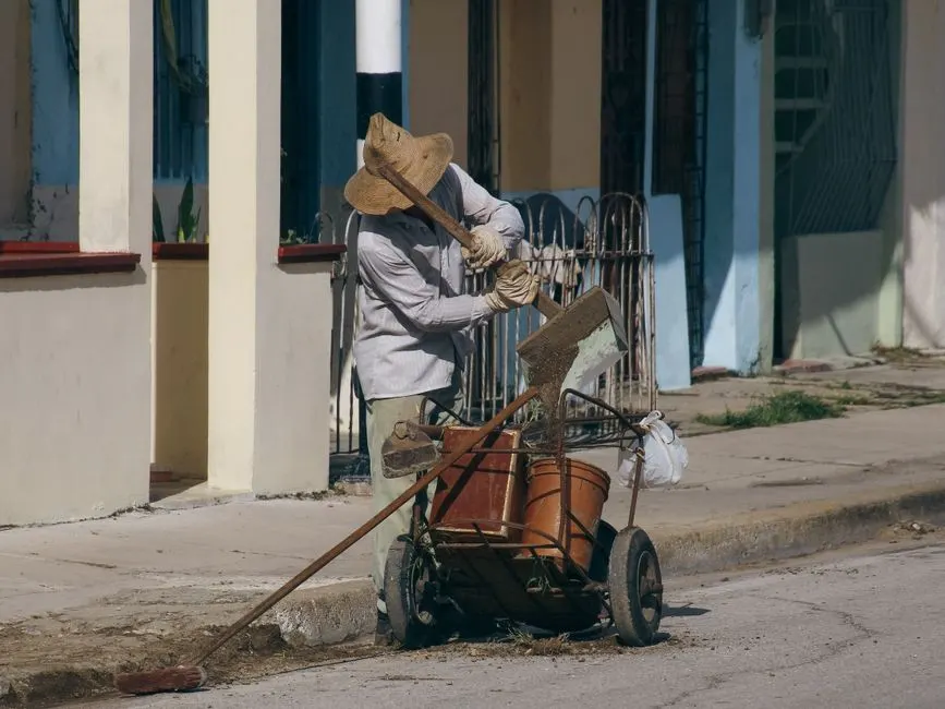 A worker in a hat sweeps and cleans a city sidewalk with a cart and broom during the day.