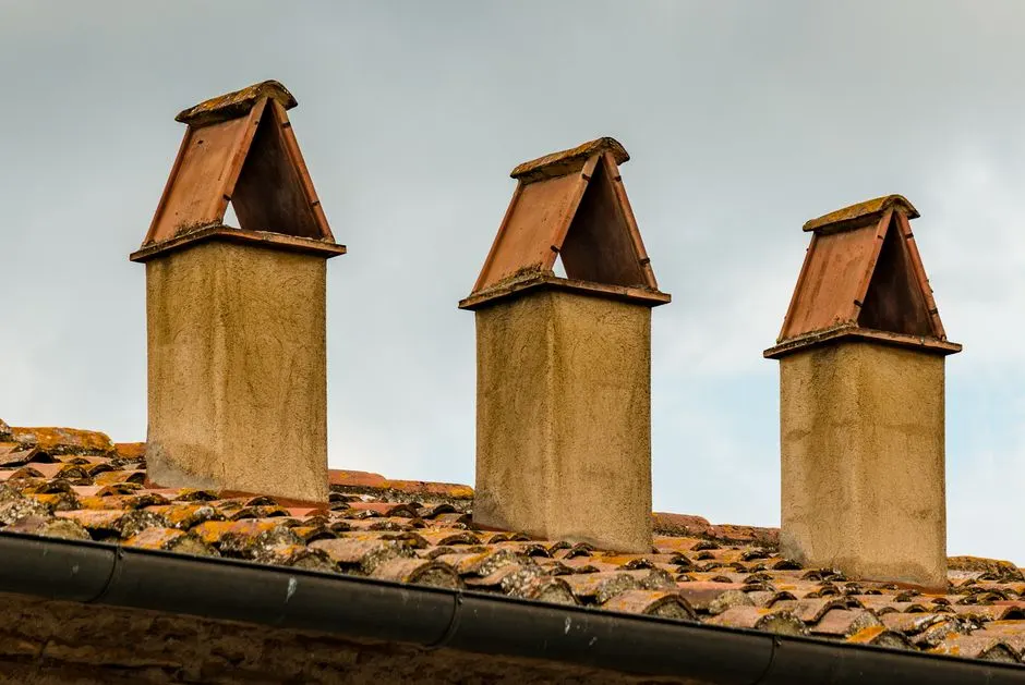 Image of three chimneys with triangular covers on a tiled roof under an overcast sky.